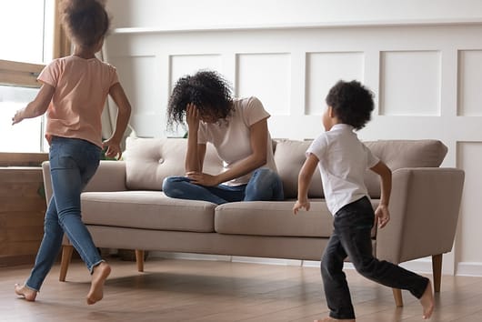 In living room mixed-race mother sit on couch cover face with hand feels desperate unhappy can't handle with noisy kids running near and make noise, misbehave children, exhausted mom need rest concept